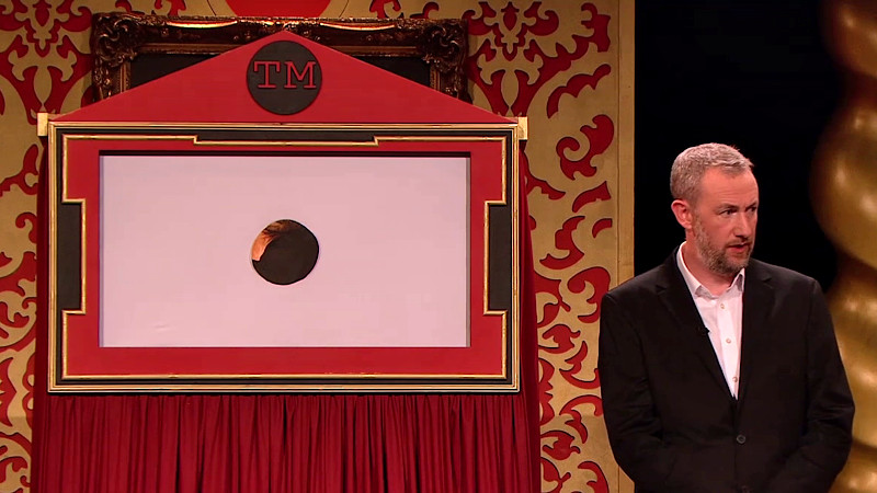 Alex Horne standing on the studio stage next to a small theatre screen with a hole in the centre. Alex Horne standing on the studio stage next to a small theatre screen with a hole in the centre.