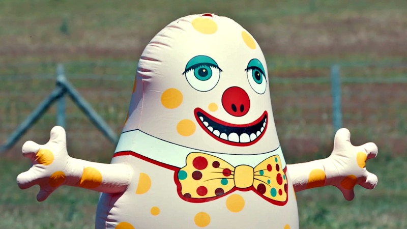 The top half of an inflatable Mr. Blobby with arms outstretched in a field. In the middle distance is an out-of-focus wire fence. The top half of an inflatable Mr. Blobby with arms outstretched in a field. In the middle distance is an out-of-focus wire fence.