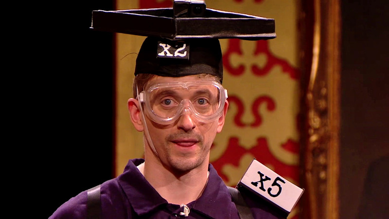 A close-up of John Robins from the shoulders up. He is wearing a black mortarboard hat with a black tray attached. It's labelled 'X2'. A white box on his shoulder reads 'X5'. He is also wearing clear safety goggles. A close-up of John Robins from the shoulders up. He is wearing a black mortarboard hat with a black tray attached. It's labelled 'X2'. A white box on his shoulder reads 'X5'. He is also wearing clear safety goggles.