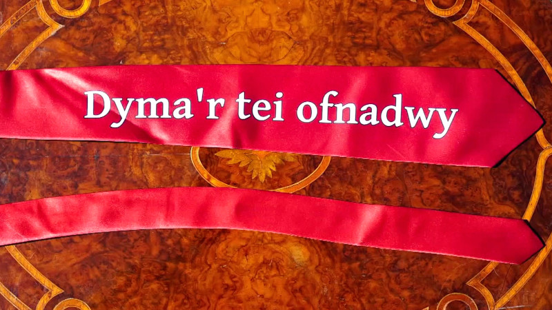 A red tie lying flat on an ornate wooden surface. On the tie is written in white the Welsh phrase 'Dyma’r tei ofnadwy' which translates to 'This is the terrible tie'. A red tie lying flat on an ornate wooden surface. On the tie is written in white the Welsh phrase 'Dyma’r tei ofnadwy' which translates to 'This is the terrible tie'.