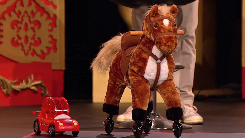 A brown and white children's toy riding horse on wheels. It's on the Taskmaster studio stage. Next to it is a small red plastic hamster wheel car. John Robins' lower body can be seen behind these toys. A brown and white children's toy riding horse on wheels. It's on the Taskmaster studio stage. Next to it is a small red plastic hamster wheel car. John Robins' lower body can be seen behind these toys.
