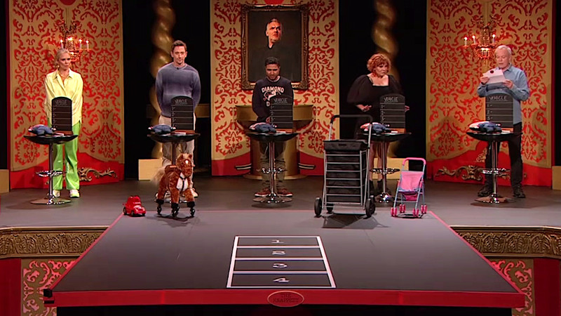 The contestants standing on the Taskmaster studio stage in front of the Knappett. Each contestant behind a table upon which there is a small stuffed doll with their own face printed on it, a small flat marker placard with their face printed on it, a small chalkboard with four lines drawn on it, and a chalk marker pen. In front of them is a brown and white children's toy riding horse on wheels. It's on the Taskmaster studio stage. Next to it is a small red plastic hamster wheel car. The contestants standing on the Taskmaster studio stage in front of the Knappett. Each contestant behind a table upon which there is a small stuffed doll with their own face printed on it, a small flat marker placard with their face printed on it, a small chalkboard with four lines drawn on it, and a chalk marker pen. In front of them is a brown and white children's toy riding horse on wheels. It's on the Taskmaster studio stage. Next to it is a small red plastic hamster wheel car.