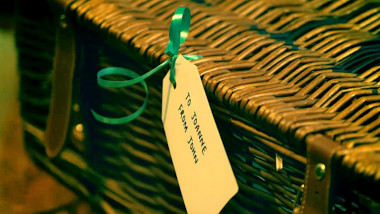 A closeup of a wicker hamper with leather straps. A gift card is attached to the hamper. It reads 'To Joanne. From John.'