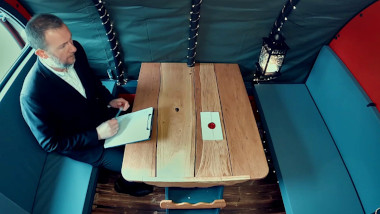 An overhead shot of Alex Horne sitting at a table in the hutch. He is holding a clipboard. On the table is a sealed task.