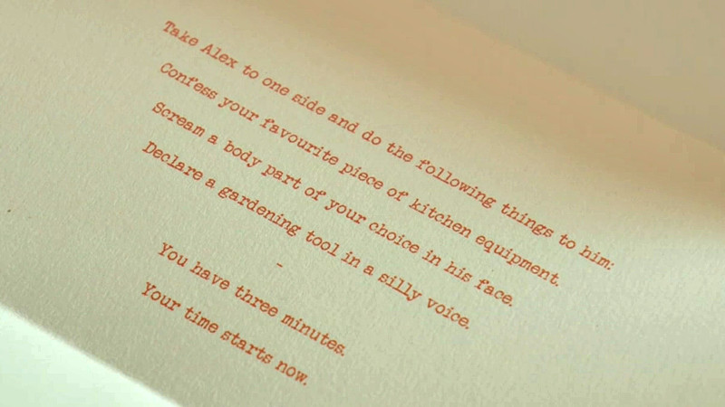 A close-up of a task. Written on the cream card in orange print the task reads 'Take Alex to one side, and do the following things to him: Confess your favourite piece of kitchen equipment. Scream a body part of your choice in his face. Declare a gardening tool in a silly voice. You have three minutes. Your time starts now.' A close-up of a task. Written on the cream card in orange print the task reads 'Take Alex to one side, and do the following things to him: Confess your favourite piece of kitchen equipment. Scream a body part of your choice in his face. Declare a gardening tool in a silly voice. You have three minutes. Your time starts now.'