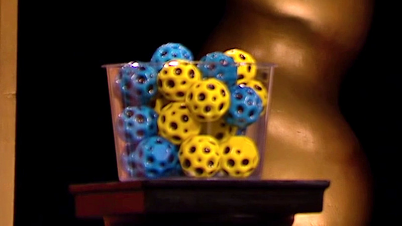 A clear plastic container on a black plinth. It is filled with yellow and blue hex balls with holes. A clear plastic container on a black plinth. It is filled with yellow and blue hex balls with holes.