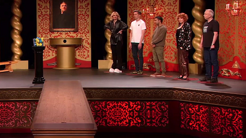 The contestants are lines up on the Taskmaster studio stage. A wooden bar extends from the front of the stage. Next to the bar is a clear plastic container on a black plinth. It is filled with yellow and blue hex balls with holes. The contestants are lines up on the Taskmaster studio stage. A wooden bar extends from the front of the stage. Next to the bar is a clear plastic container on a black plinth. It is filled with yellow and blue hex balls with holes.