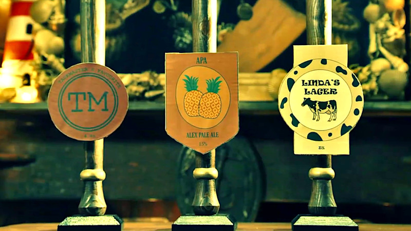 Three taps; one with the Taskmaster logo and the words 'Taskmaster & Favourite' on it, one with pineapples on the front titled 'Alex Pale Ale', and one with a cow and cow print titled 'Linda's Lager'. Three taps; one with the Taskmaster logo and the words 'Taskmaster & Favourite' on it, one with pineapples on the front titled 'Alex Pale Ale', and one with a cow and cow print titled 'Linda's Lager'.