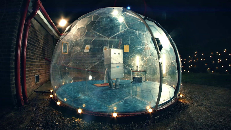 A robot inside the geodesic dome, with caution signs, a list, and a painting around. A doormat sits behind the robot, red wine on a table with a safe underneath sits on the left of it, and a blue siren is put on top of the dome. A robot inside the geodesic dome, with caution signs, a list, and a painting around. A doormat sits behind the robot, red wine on a table with a safe underneath sits on the left of it, and a blue siren is put on top of the dome.