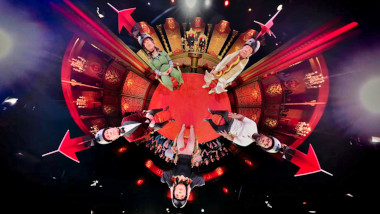 A 360-degree shot of all five contestants sitting in a circle and wearing helmets with large red arrows.